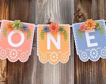 May include: A colorful paper banner with the word "ONE" in white letters, decorated with paper flowers in shades of orange, blue, and green. The banner is strung on a white ribbon and has a scalloped edge.