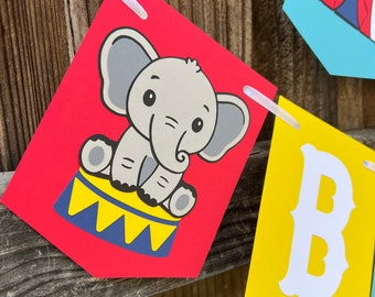 May include: A colorful circus-themed banner with pennants. One pennant features a cartoon elephant on a circus drum against a red background. Other pennants have a circus tent and the letter "B". The banner is strung on a white ribbon.