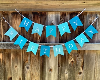 May include: A blue and white banner with the words "Happy Birthday" strung on a white string. The banner is hanging on a wooden fence.