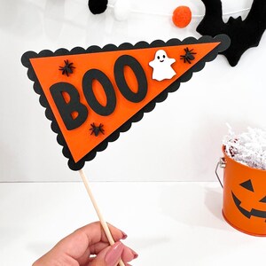 May include: Orange and black Halloween party flag with the word "BOO" in black letters and a white ghost. The flag is attached to a wooden stick.