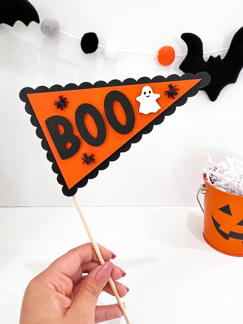 May include: Orange and black Halloween party flag with the word "BOO" in black letters and a white ghost. The flag is attached to a wooden stick.