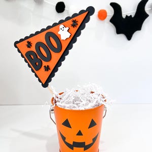 May include: Orange pumpkin-shaped bucket with a black jack-o'-lantern face filled with white shredded paper. A black and orange triangle flag with the word "BOO" in black letters and a white ghost is attached to a wooden stick.