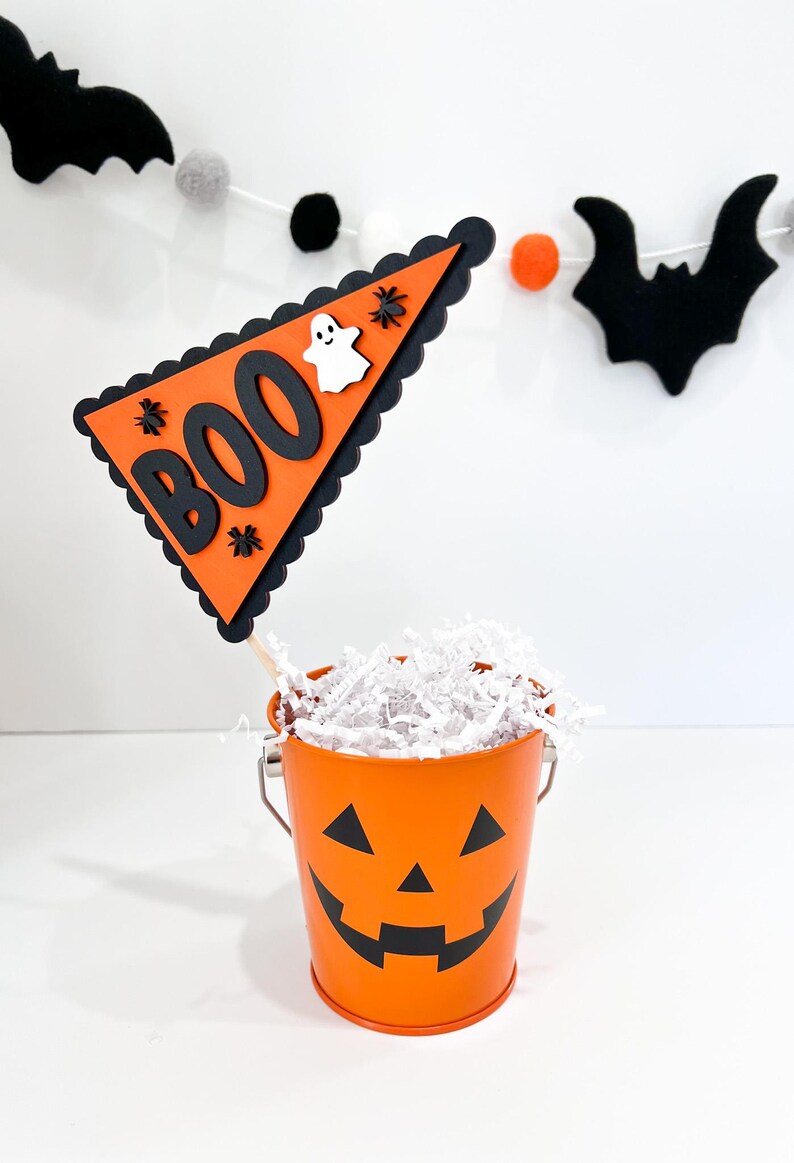 May include: Orange pumpkin-shaped bucket with a black jack-o'-lantern face filled with white shredded paper. A black and orange triangle flag with the word "BOO" in black letters and a white ghost is attached to a wooden stick.
