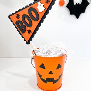 May include: Orange pumpkin-shaped metal bucket with a black jack-o'-lantern face. The bucket is filled with white shredded paper and a black and orange "BOO" pennant with a white ghost and black spiders.