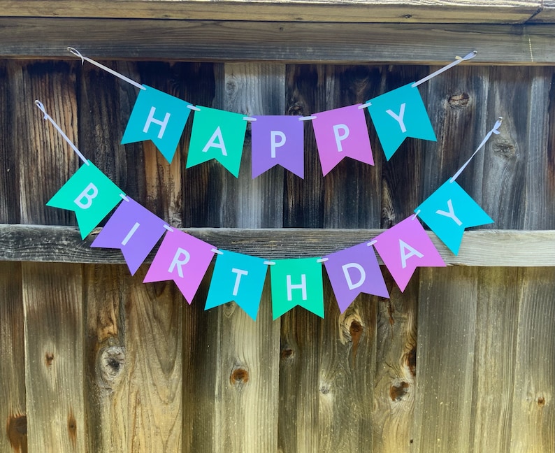 May include: A colorful banner with the words "Happy Birthday" in white letters on a teal, purple, and pink background. The banner is strung on a white string and is hanging on a wooden fence.