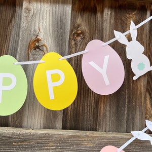 以下が含まれることがあります： 卵の形をした紙の切り抜きに「Happy」という文字が書かれた、パステルカラーのイースターバナー。白いリボンで結ばれており、白いウサギのシルエットが2つ描かれている。