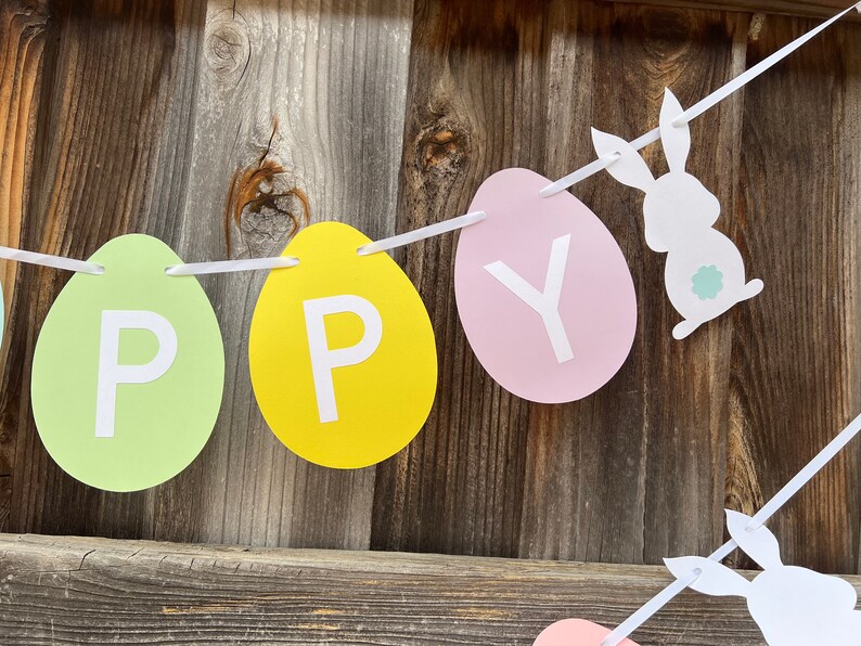以下が含まれることがあります： 卵の形をした紙の切り抜きに「Happy」という文字が書かれた、パステルカラーのイースターバナー。白いリボンで結ばれており、白いウサギのシルエットが2つ描かれている。