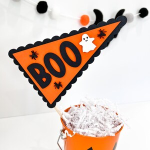 May include: Orange and black Halloween party decoration with a triangular flag that says "BOO" with a ghost and spiders. The flag is attached to a wooden stick and is in a small orange pumpkin bucket.