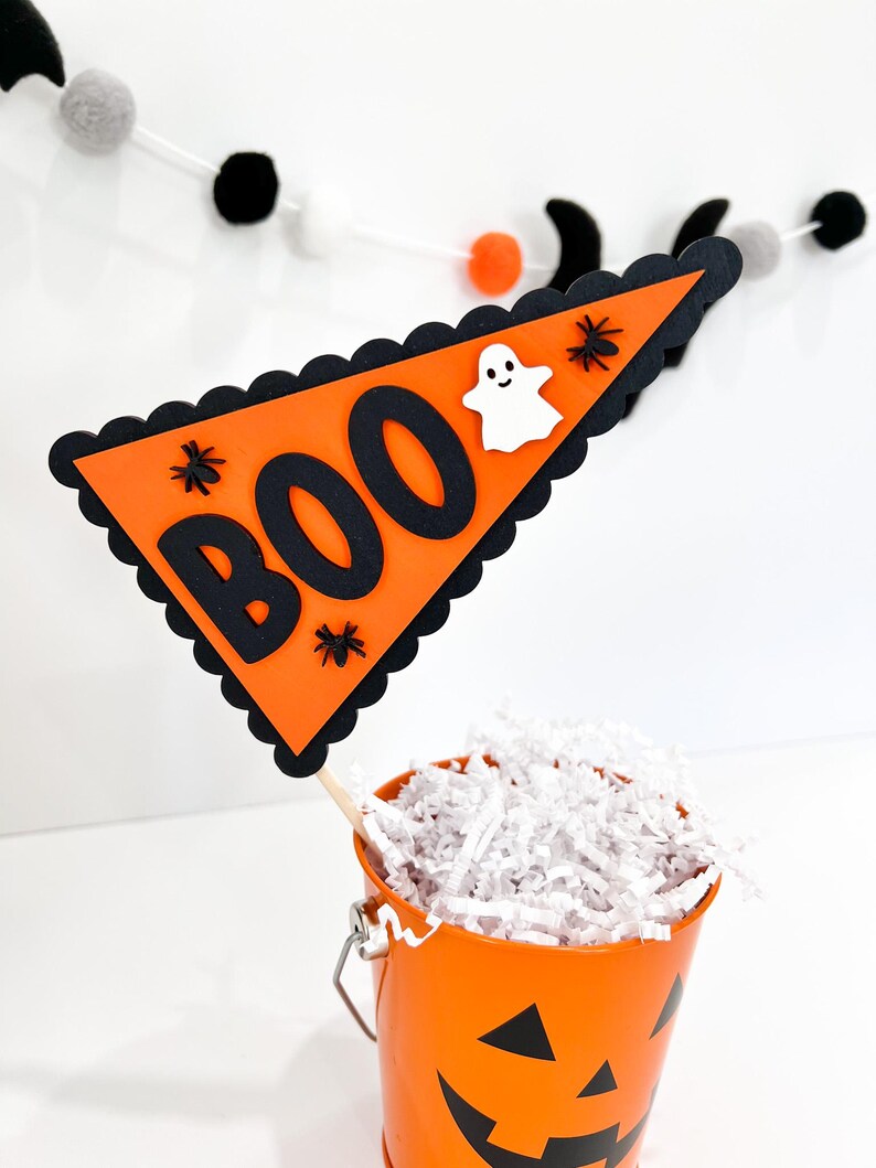 May include: Orange and black Halloween party decoration with a triangular flag that says "BOO" with a ghost and spiders. The flag is attached to a wooden stick and is in a small orange pumpkin bucket.