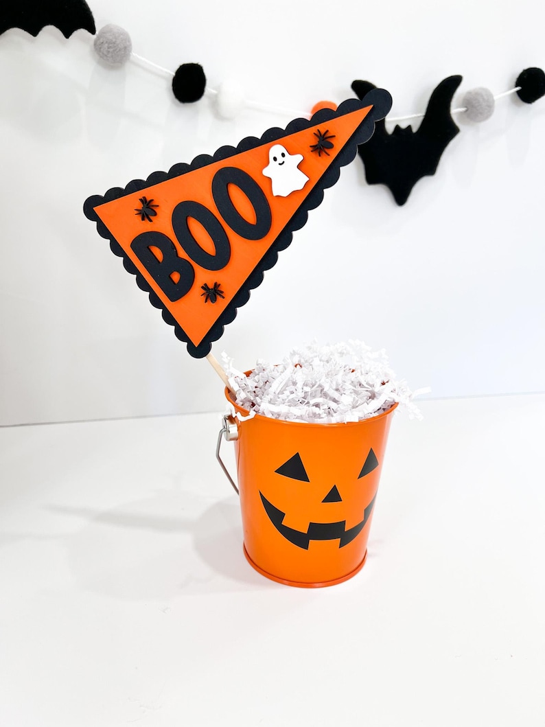 May include: Orange pumpkin-shaped bucket with a black jack-o'-lantern face. The bucket is filled with white shredded paper and a black and orange pennant flag with the word "BOO" in black letters and a white ghost.