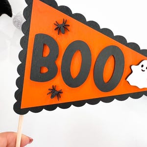 May include: Orange and black Halloween banner with the word "BOO" in black letters. The banner has a scalloped edge and is decorated with black spiders and a white ghost.