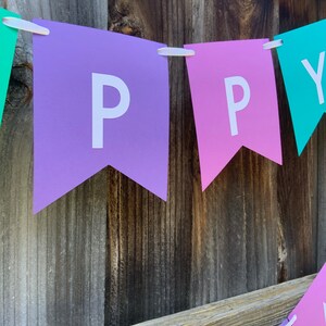 May include: A purple, pink, and teal banner with the word "HAPPY" spelled out in white letters. The banner is hanging on a wooden fence.