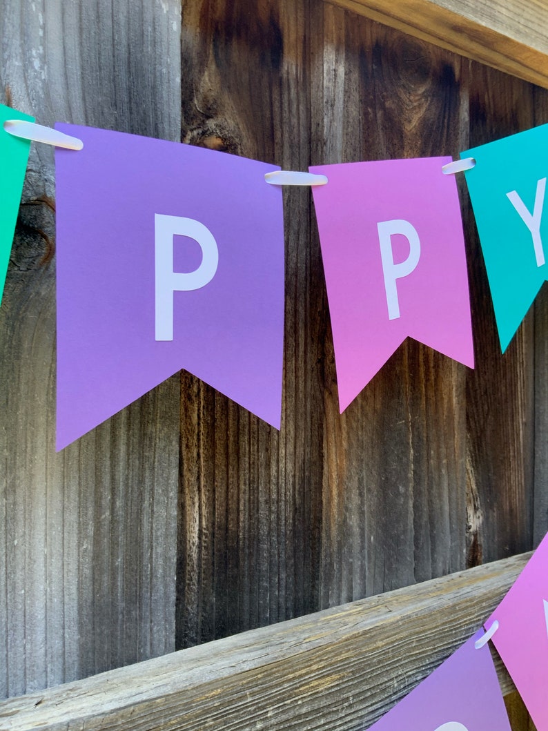 May include: A purple, pink, and teal banner with the word "HAPPY" spelled out in white letters. The banner is hanging on a wooden fence.