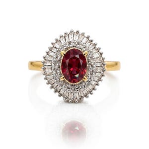 May include: A gold ring featuring a central oval-cut deep red gemstone held by four prongs. The stone is surrounded by a double halo consisting of an inner circle of small round white stones and an outer border of baguette-cut white stones.