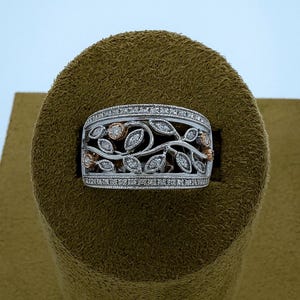 Puede incluir: Un anillo de plata con un diseño floral. El anillo presenta una banda con un patrón de filigrana de hojas y flores, acentuado con pequeños diamantes. Las flores tienen un tono oro rosa, y el anillo está sobre una superficie marrón.