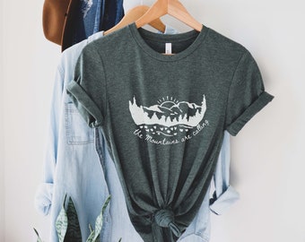 May include: A dark green t-shirt with a white graphic of mountains and a sun. The text "the mountains are calling" is printed below the graphic.