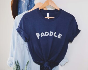 Puede incluir: Una camiseta azul marino con la palabra "PADDLE" impresa en blanco en la parte delantera. La camiseta está colgada de una percha.