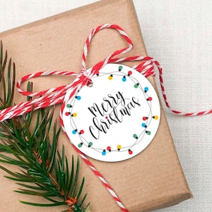 Puede incluir: Una caja de regalo marrón envuelta con hilo rojo y blanco y una etiqueta de regalo redonda con el texto "Feliz Navidad" y una cadena de luces de colores.