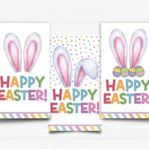 Puede incluir: Tres tarjetas imprimibles con fondo blanco y rayas de colores. Cada tarjeta presenta una ilustración de acuarela de orejas de conejo y el texto "¡Feliz Pascua!" en colores arcoíris.