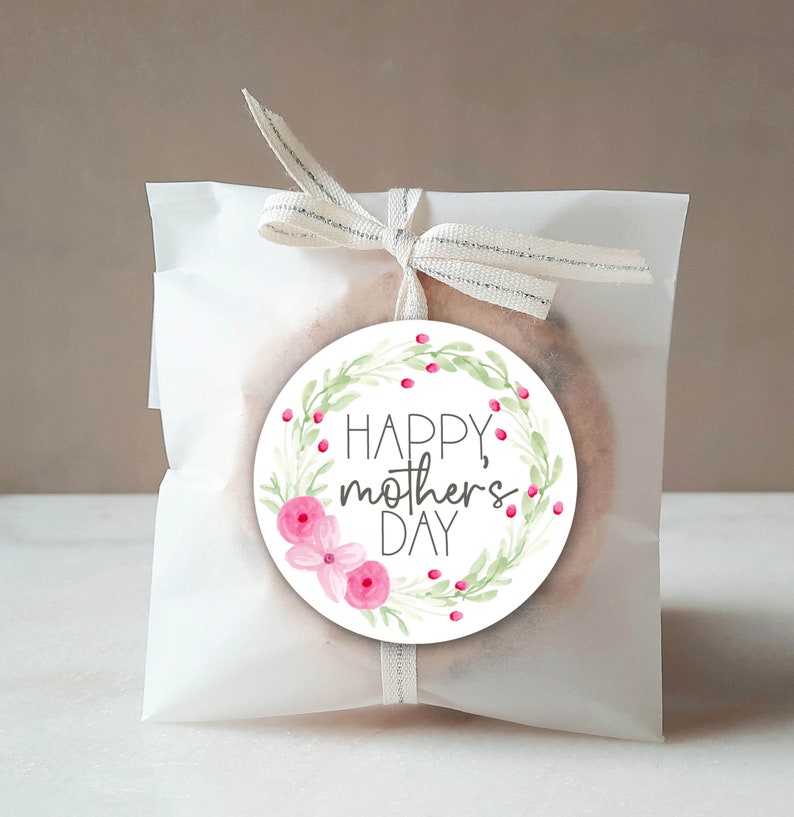 May include: A gift wrapped in white paper and tied with a ribbon. A circular sticker with the text "HAPPY, mothers DAY" is attached. The sticker features a floral design with pink flowers and green leaves.