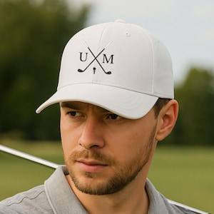 Könnte beinhalten: Eine weiße Baseballkappe mit den Buchstaben "UXM" und gekreuzten Golfschlägern in Schwarz. Die Kappe hat einen gebogenen Schirm. Die Person trägt ein graues Poloshirt.