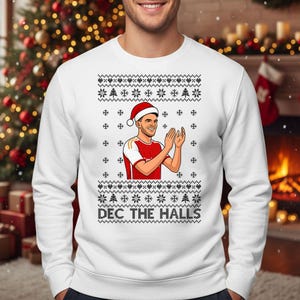 May include: White sweatshirt with a festive design. The graphic features a person wearing a Santa hat, clapping, and wearing a red and white jersey. The text "DEC THE HALLS" is printed below the graphic. The sweatshirt has a snowflake and Christmas tree pattern.