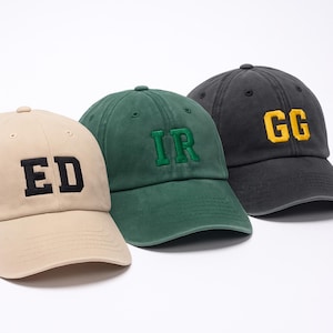 Könnte beinhalten: Drei Baseballkappen mit gestickten Buchstaben. Die erste ist beige mit schwarzen Buchstaben "ED", die zweite ist grün mit grünen Buchstaben "IR" und die dritte ist dunkelgrau mit gelben Buchstaben "GG".