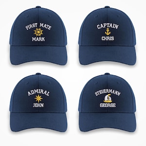 May include: Four navy blue baseball caps with white embroidered text and nautical designs. Each cap displays a different title: "First Mate," "Captain," "Admiral," and "Steuermann," along with a name. The designs include a ship's wheel, anchor, starburst, and sailboat.