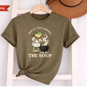 May include: Khaki green t-shirt with a cartoon frog chef and the text "That's it. You're going in THE SOUP." The frog is wearing a chef's hat and apron, stirring a pot. The shirt is hanging on a wooden hanger.