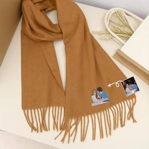 May include: A tan scarf with a fringed edge. The scarf has a small illustration of a couple and a photo attached. The scarf is made of a soft material and is folded on a white surface.