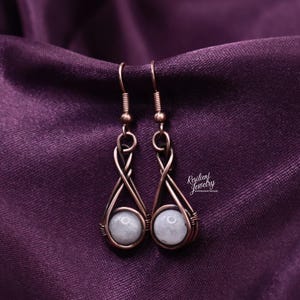 May include: Copper-colored wire earrings with a teardrop design, each holding a round, pale blue-gray stone. The earrings hang from a simple hook and are displayed against a deep purple fabric. The earrings are from Resilient Jewelry.