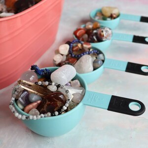 May include: Five turquoise measuring cups filled with various colorful gemstones and crystals. The cups have black handles and are arranged in a row on a white surface.