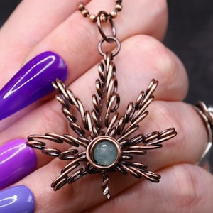 Könnte beinhalten: Kupferfarbene Anhängerkette mit einem Cannabisblatt-Design. Das Blatt ist aus gedrehtem Draht gefertigt, mit einem kleinen, runden, hellgrünen Stein in der Mitte. Die Halskette hat eine Kugelkette.