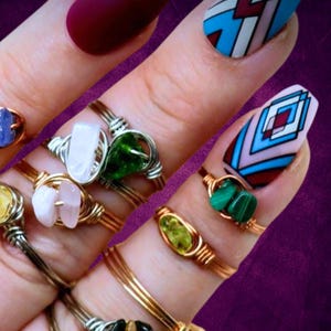 May include: A hand wearing several rings with different colored stones wrapped in wire. The rings are made of gold and silver wire. The stones are a variety of colors, including blue, green, yellow, and pink. The hand is also wearing a red nail polish on one finger and a blue and white geometric design on another finger.