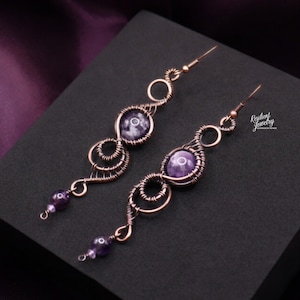 May include: A pair of handcrafted copper wire earrings with purple amethyst beads and stones. The earrings feature a spiral design with a hook closure. The earrings are displayed on a black surface with a purple satin background.