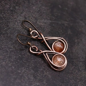May include: A pair of copper wire earrings with a sunstone bead on each earring. The earrings are in the shape of a teardrop with a swirl design.