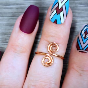 Puede incluir: Una mano con un anillo de oro con un diseño de doble espiral. El anillo está en el dedo medio. Las uñas están pintadas con un esmalte burdeos mate y un diseño geométrico azul y blanco.