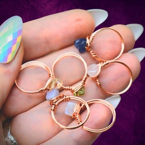 Può includere: Una collezione di anelli in filo di rame, ognuno adornato con una gemma diversa. Le gemme includono pietre blu, verdi, gialle e trasparenti. Gli anelli sono presentati su uno sfondo viola.