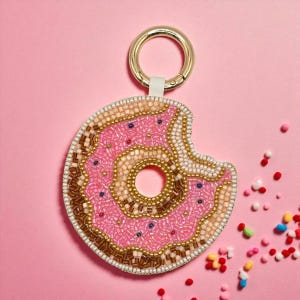 May include: A pink beaded donut keyring with a bite taken out, featuring brown, gold, and white accents. The keyring has a gold ring and a white strap. Colourful sprinkles are scattered on a pink background.