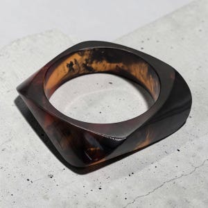May include: A square bangle bracelet in shades of brown and amber. The bracelet has a glossy finish and a marbled appearance, with a geometric design. The bracelet is made of a solid material.
