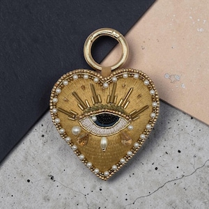 Puede incluir: Un llavero en forma de corazón con bordados y abalorios en tono dorado. El centro muestra un motivo de ojo con pupila oscura, iris blanco y rayos dorados. El borde está delineado con pequeñas perlas blancas y tiene una argolla de metal dorado.