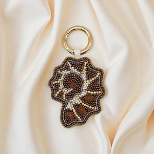 May include: A beaded keychain with a gold-coloured clasp. The pendant is brown with white, gold, and brown beadwork in a starburst design, and features several brown gemstone accents. The keychain is set against a cream-coloured fabric.