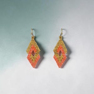 Könnte beinhalten: Ein Paar handgefertigte Perlenohrringe. Jeder Ohrring hat eine rautenförmige Form mit goldener Ober- und Unterseite und einer korallenfarbenen Mitte. Die Ohrringe hängen an goldfarbenen Ohrhaken.