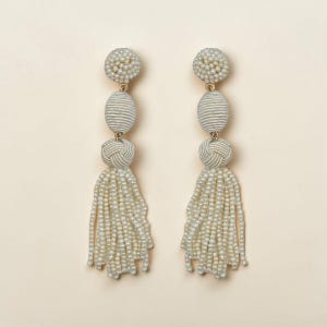 May include: A pair of off-white tassel earrings. Each earring has a beaded top, a ribbed oval bead, a knotted bead, and a long tassel of small, dangling beads. The earrings have gold-coloured metal connecting hardware.