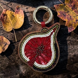 May include: A fig-shaped keyring featuring intricate beadwork and sequins in shades of deep red, white, and green. The design mimics the cross-section of a sliced fig, with a gold-toned metal ring attached to the top by a small red leather tab.