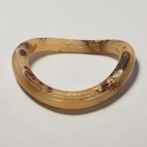 Op de afbeelding: Een lichtbruine, ovaalvormige armband met donkerbruine en crèmekleurige marmering. De armband is gemaakt van een doorschijnend materiaal en heeft een gladde, gepolijste afwerking. Het ontwerp is eenvoudig en elegant.