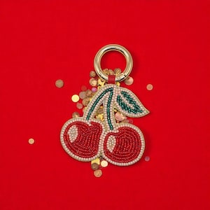Op de afbeelding: Een sleutelhanger in de vorm van een kers, met rode, met kralen versierde kersen, groene bladeren en een goudkleurige ring. De kersen zijn gedetailleerd met rode en witte kralen. De achtergrond is effen rood met gouden confetti.