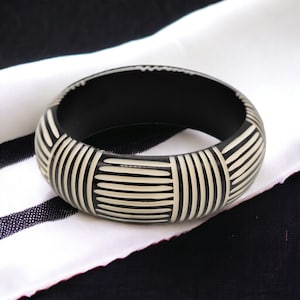 Könnte beinhalten: Schwarz-weißes Armreif-Armband mit geometrischem Muster. Das Armband ist aus einem glatten, harten Material gefertigt.