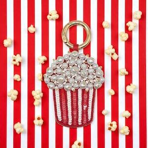 Puede incluir: Un llavero con forma de palomitas de maíz con un anillo dorado y una correa roja. La parte superior está adornada con perlas y cuentas blancas, mientras que la base presenta lentejuelas rojas y blancas. El fondo es un patrón de rayas rojas y blancas, con palomitas de maíz dispersas.