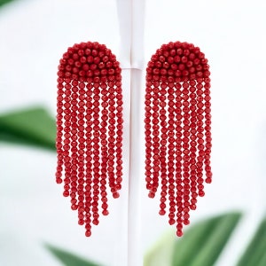 May include: A pair of long red beaded earrings with a round top. The earrings are made of small red beads that are strung together in a cascading pattern.